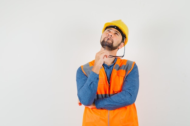 Giovane costruttore in camicia, gilet, casco mordere gli occhiali, alzando lo sguardo e guardando pensieroso, vista frontale.