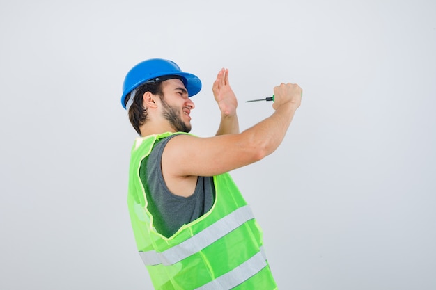 Giovane costruttore in uniforme che finge di colpirsi con un cacciavite e sembra pazzo, vista frontale.