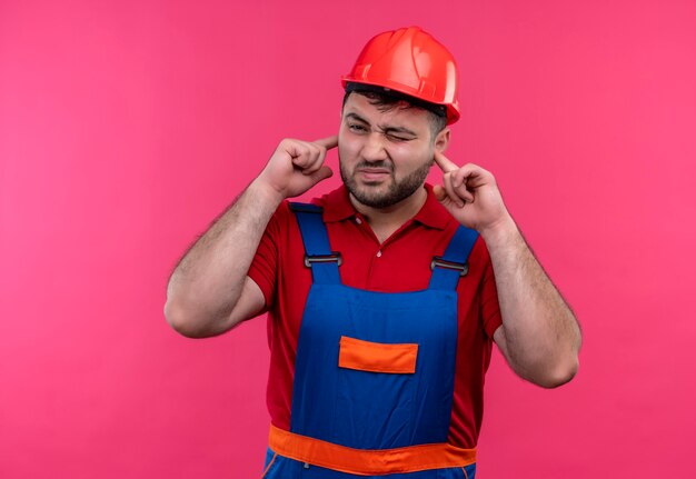 Giovane costruttore in uniforme da costruzione e casco di sicurezza che copre le orecchie con le dita con espressione infastidita per il rumore del suono forte