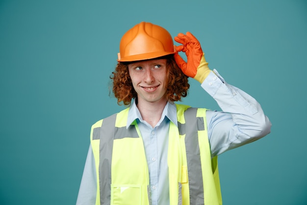 Giovane costruttore in uniforme da costruzione e casco di sicurezza che indossa guanti di gomma che guarda da parte sorridente allegramente felice e positivo in piedi su sfondo blu