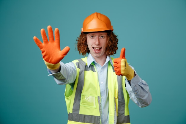 Giovane costruttore in uniforme da costruzione e casco di sicurezza che indossa guanti di gomma guardando la fotocamera che mostra il numero cinque con il palmo aperto che mostra il pollice in alto sorridente in piedi su sfondo blu