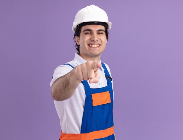 Giovane costruttore in uniforme da costruzione e casco di sicurezza che punta con il dito indice davanti sorridendo allegramente in piedi sopra la parete viola