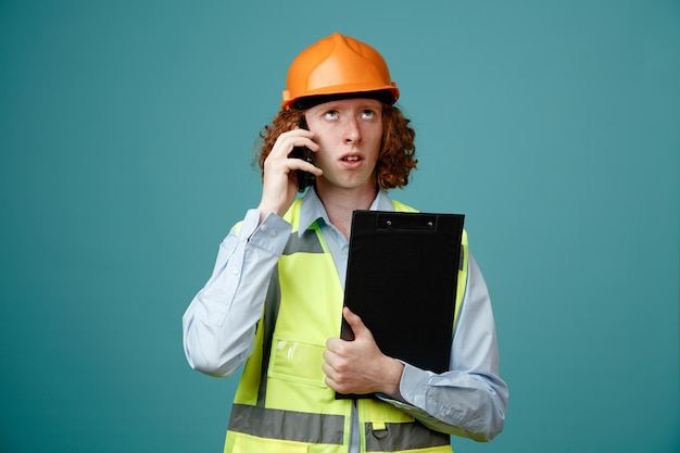 Giovane costruttore in uniforme da costruzione e casco di sicurezza che tiene appunti parlando sul telefono cellulare cercando confuso in piedi su sfondo blu