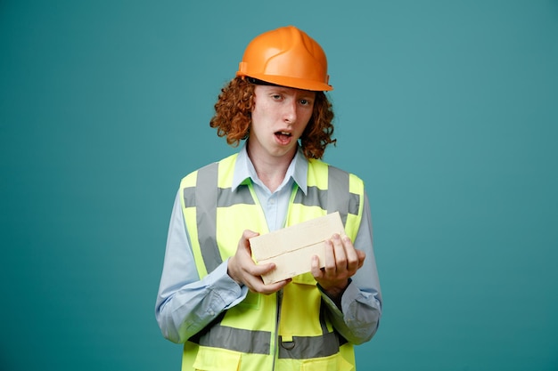 Giovane costruttore in uniforme da costruzione e casco di sicurezza che tiene due mattoni guardando la fotocamera confuso in piedi su sfondo blu