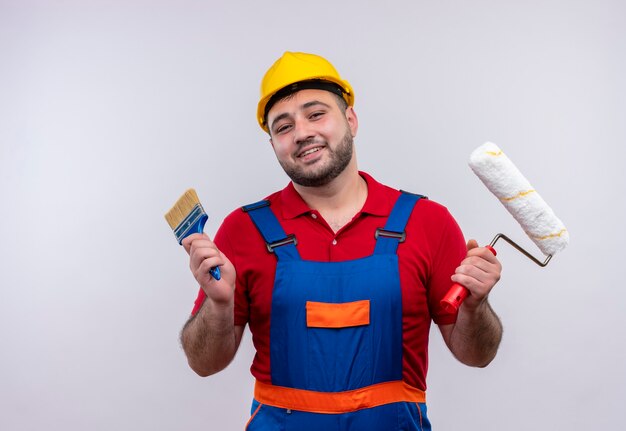 Giovane costruttore in uniforme da costruzione e casco di sicurezza che tiene il rullo della pinta e la spazzola sorridendo allegramente