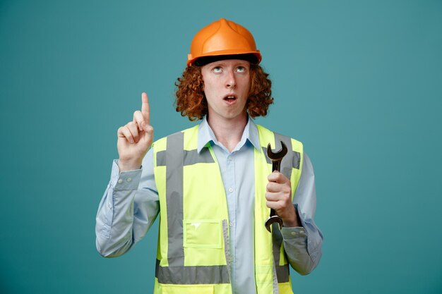 Giovane costruttore in uniforme da costruzione e casco di sicurezza che tiene la chiave che punta con il dito indice verso l'alto guardando sorpreso in piedi su sfondo blu