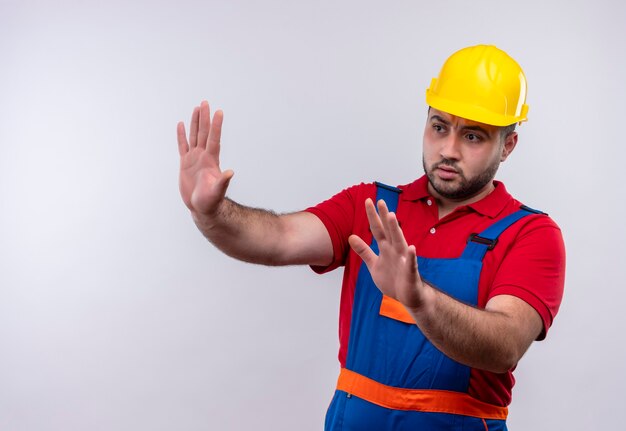 Giovane costruttore in uniforme da costruzione e casco di sicurezza che tiene le mani in alto facendo gesto di difesa con espressione di paura