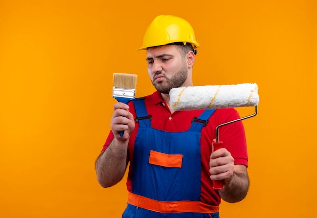 Giovane costruttore in uniforme da costruzione e casco di sicurezza che tiene pennello e rullo di vernice guardando il pennello con espressione scettica