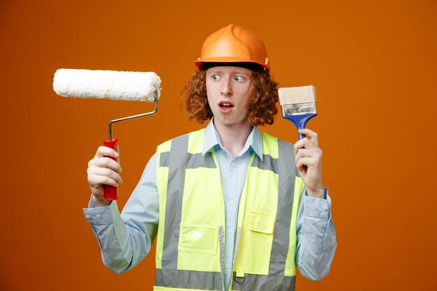 Giovane costruttore in uniforme da costruzione e casco di sicurezza che tiene rullo di vernice e pennello che sembra confuso avendo dubbi in piedi su sfondo arancione