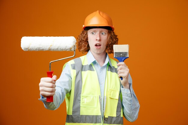 Giovane costruttore in uniforme da costruzione e casco di sicurezza che tiene rullo di vernice e pennello che sembra confuso avendo dubbi in piedi su sfondo arancione