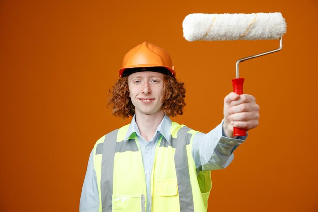 Giovane costruttore in uniforme da costruzione e casco di sicurezza che tiene rullo di vernice guardando la fotocamera sorridente fiducioso felice e positivo in piedi su sfondo arancione