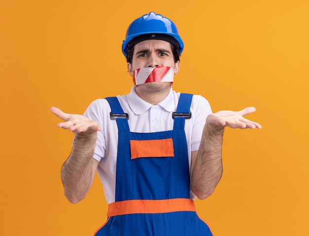 Giovane costruttore in uniforme da costruzione e casco di sicurezza con nastro adesivo avvolto intorno alla bocca che guarda confuso scrollare le spalle in piedi sopra la parete arancione