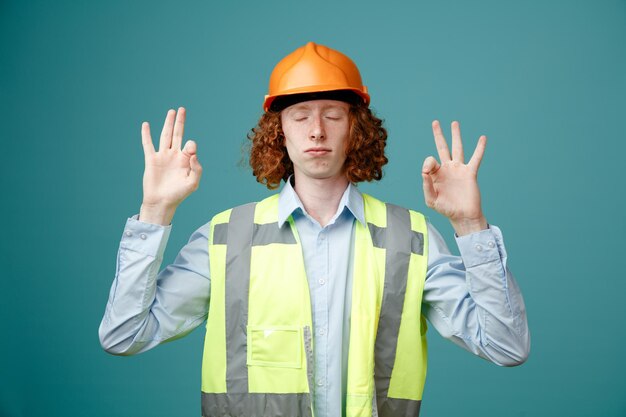 Giovane costruttore in uniforme da costruzione e casco di sicurezza meditando rilassante facendo gesto di meditazione in piedi su sfondo blu