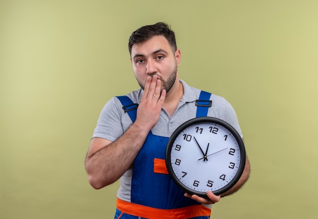 Giovane costruttore in uniforme di costruzione che tiene l'orologio da parete scioccato che copre la bocca con la mano