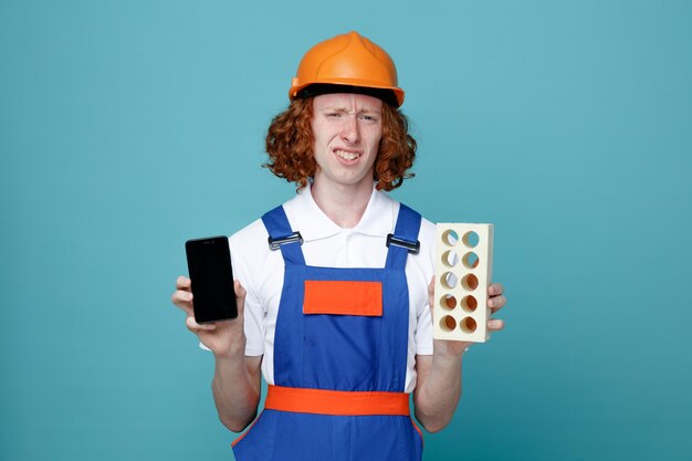 giovane costruttore scontento in uniforme che tiene il telefono con mattoni isolati su sfondo blu