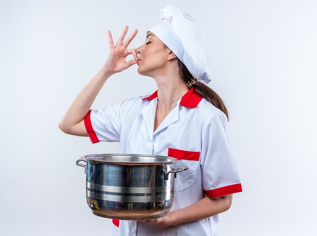 Giovane cuoca che indossa l'uniforme dello chef tenendo la casseruola isolata sulla parete bianca?