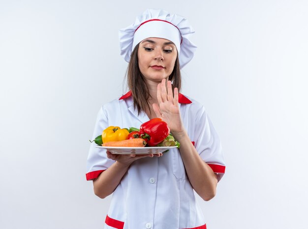 Giovane cuoca scontenta che indossa l'uniforme da chef che tiene le verdure sulla piastra che mostra il gesto di arresto isolato su sfondo bianco