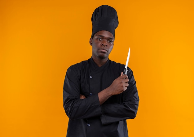 Giovane cuoco afroamericano fiducioso in uniforme da chef incrocia le braccia e tiene il coltello che guarda l'obbiettivo sull'arancio con lo spazio della copia
