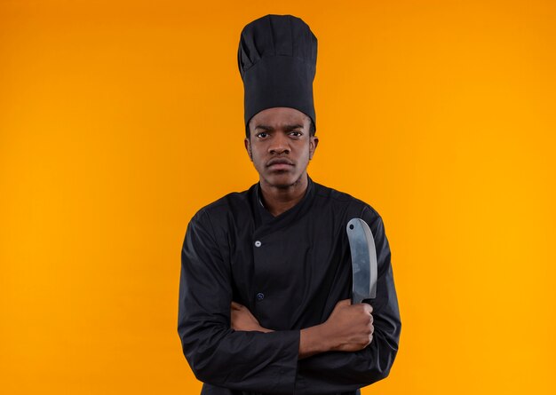 Giovane cuoco afroamericano fiducioso in uniforme da chef incrocia le braccia e tiene il coltello isolato su sfondo arancione con spazio di copia