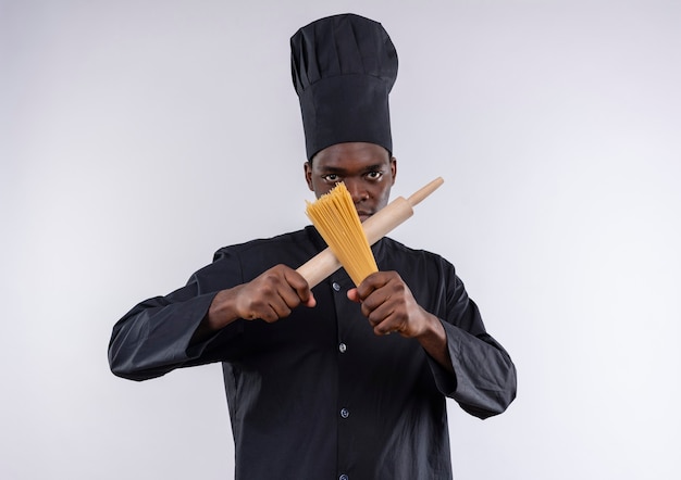 Giovane cuoco afroamericano sicuro in uniforme del cuoco unico attraversa il mattarello e il mazzo di spaghetti su bianco con lo spazio della copia