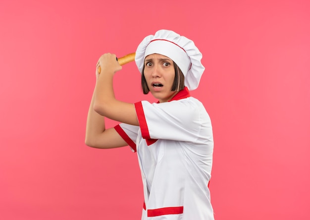 Giovane cuoco arrabbiato femminile in uniforme del cuoco unico che tiene il grissino e si prepara a battere qualcuno isolato sul rosa con lo spazio della copia