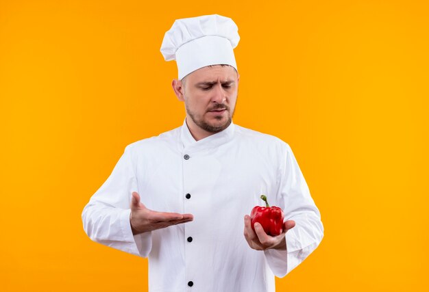 Giovane cuoco bello dispiaciuto in uniforme del cuoco unico che tiene che osserva e che indica al pepe isolato sullo spazio arancione