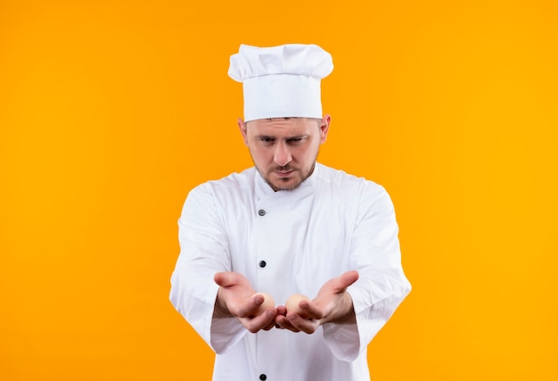 Giovane cuoco bello in uniforme del cuoco unico che allunga e che mostra le mani vuote e li guarda isolati sullo spazio arancione