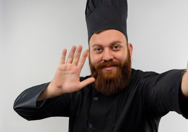 Giovane cuoco bello in uniforme del cuoco unico che gesturing ciao e che allunga la mano isolata su spazio bianco