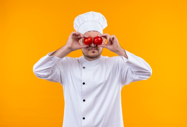 Giovane cuoco bello in uniforme del cuoco unico che mette i pomodori sugli occhi isolati sullo spazio arancione