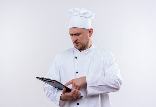 Giovane cuoco bello in uniforme del cuoco unico che tiene, che osserva e che mette il dito negli appunti isolato su spazio bianco