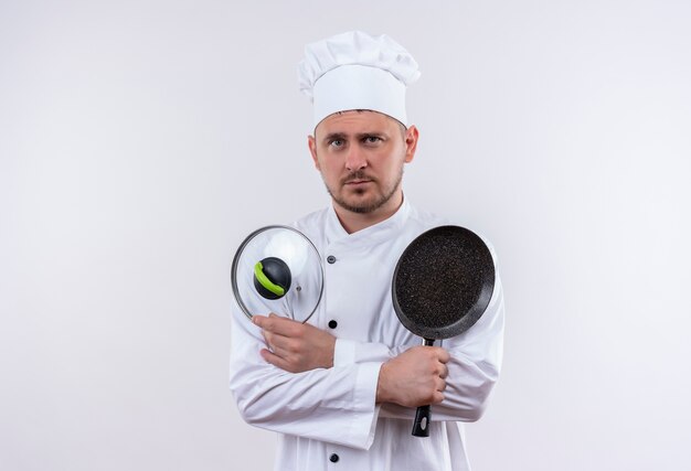 Giovane cuoco bello in uniforme del cuoco unico che tiene la padella e il coperchio della padella che sembrano isolati su spazio bianco