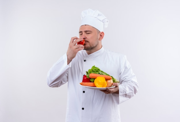 Giovane cuoco bello in uniforme del cuoco unico che tiene piatto con verdure e annusando il pomodoro con gli occhi chiusi su uno spazio bianco isolato