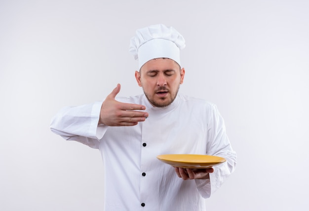 Giovane cuoco bello nel piatto della tenuta dell'uniforme del cuoco unico e annusando con la mano alzata e gli occhi chiusi isolati su spazio bianco