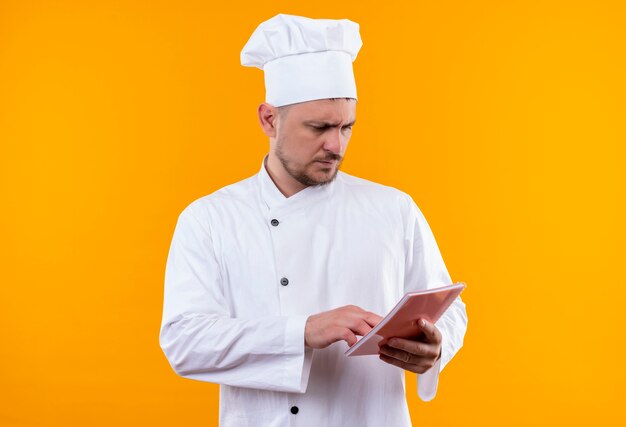 Giovane cuoco bello premuroso in rilievo di nota della tenuta dell'uniforme del cuoco unico che mette la mano sopra e che lo esamina isolato sullo spazio arancione