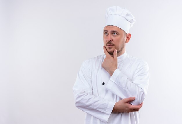 Giovane cuoco bello premuroso in uniforme del cuoco unico che mette la mano sul mento che esamina il lato isolato su spazio bianco