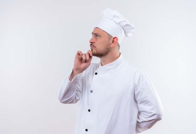 Giovane cuoco bello premuroso in uniforme del cuoco unico che mette la mano sul mento che sta nella vista di profilo che esamina il lato isolato su spazio bianco