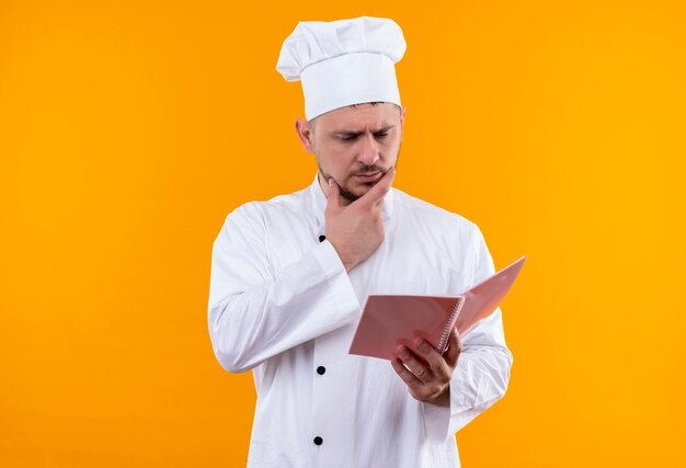 Giovane cuoco bello premuroso in uniforme del cuoco unico che tiene e che esamina il rilievo di nota con la mano sul mento isolato sullo spazio arancione