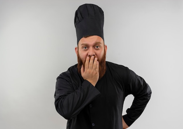 Giovane cuoco bello sorpreso in uniforme del cuoco unico che mette la mano sulla bocca con la mano sulla vita isolata su spazio bianco