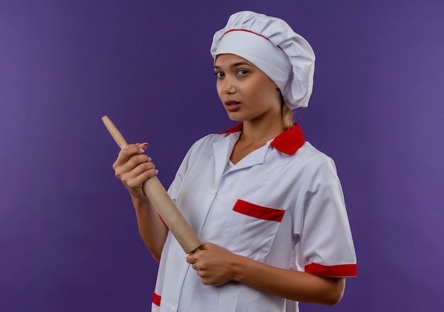 giovane cuoco femmina che indossa uniforme chef tenendo il mattarello sulla parete isolata