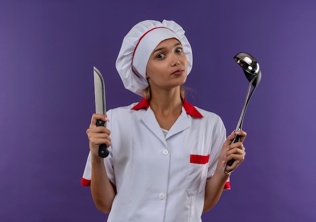 giovane cuoco femmina che indossa uniforme chef tenendo mestolo e coltello sulla parete isolata