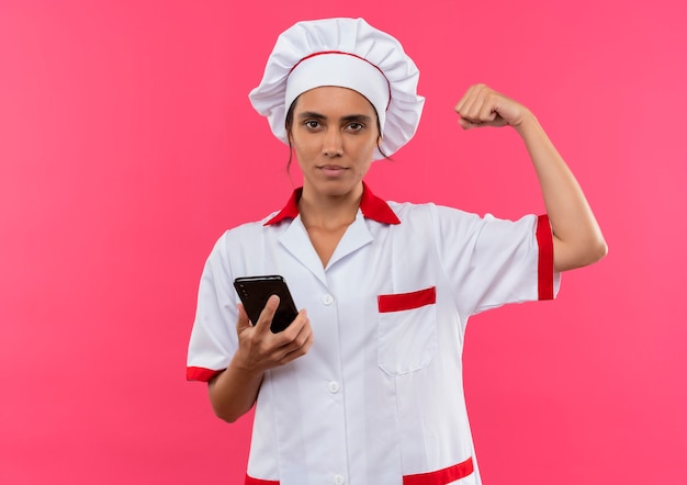 giovane cuoco femmina che indossa uniforme da chef tenendo il telefono e facendo un forte gesto sulla parete rosa isolata con lo spazio della copia