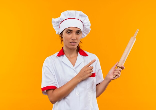 giovane cuoco femminile che indossa la tenuta uniforme dello chef e indica il mattarello sulla parete gialla isolata con spazio di copia