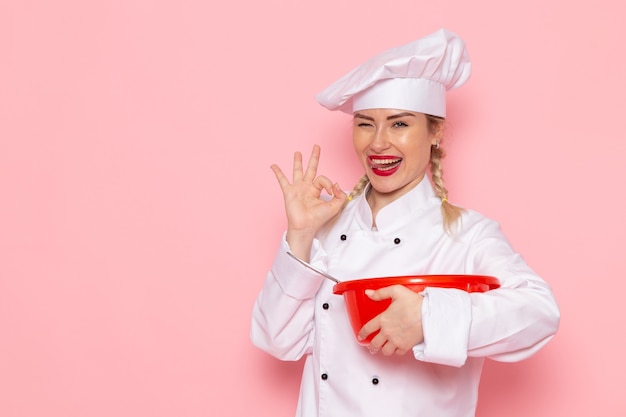 Giovane cuoco femminile di vista frontale in vestito bianco che tiene ciotola rossa che sorride sulla foto del lavoro di lavoro di cucina del cuoco dello spazio rosa