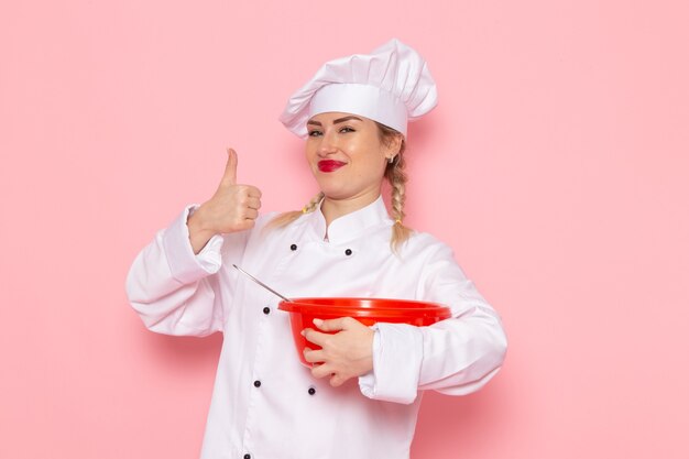 Giovane cuoco femminile di vista frontale in vestito bianco che tiene ciotola rossa che sorride sulla foto del lavoro di lavoro di cucina del cuoco dello spazio rosa