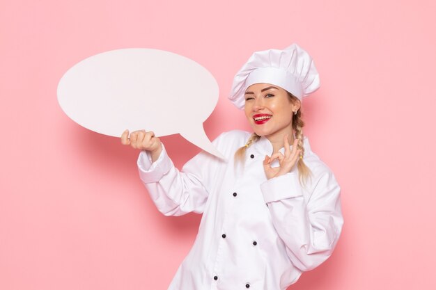 Giovane cuoco femminile di vista frontale in vestito bianco del cuoco che tiene qualche segno bianco che sorride sul cuoco rosa dello spazio
