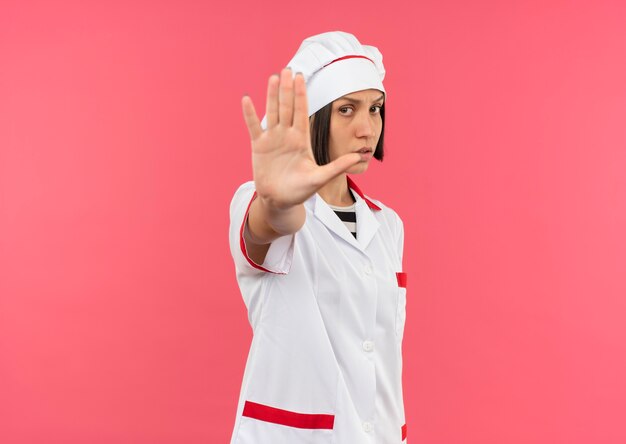 Giovane cuoco femminile insoddisfatto in uniforme del cuoco unico che gesturing fermata alla macchina fotografica isolata su fondo rosa con lo spazio della copia