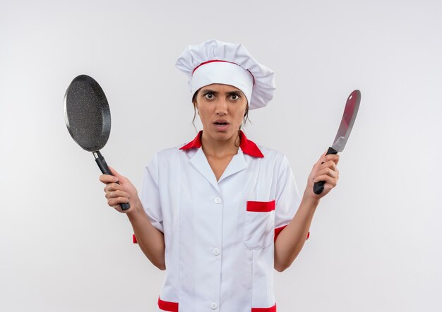 Giovane cuoco femminile interessato che indossa l'uniforme del cuoco unico che tiene la mannaia e la padella sulla parete bianca isolata con lo spazio della copia