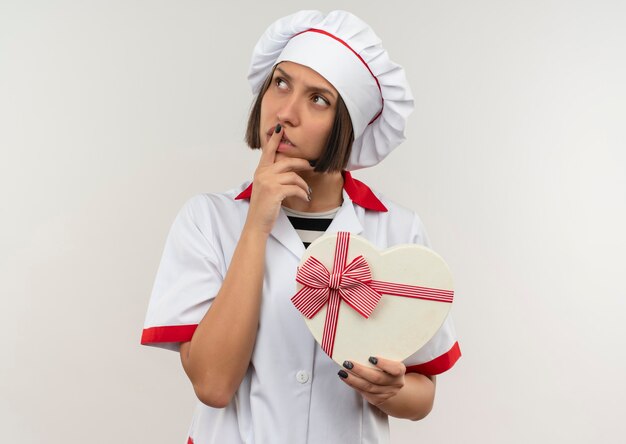 Giovane cuoco femminile premuroso in uniforme del cuoco unico che tiene il contenitore di regalo a forma di cuore che esamina lato con il dito sulle labbra isolate sulla parete bianca