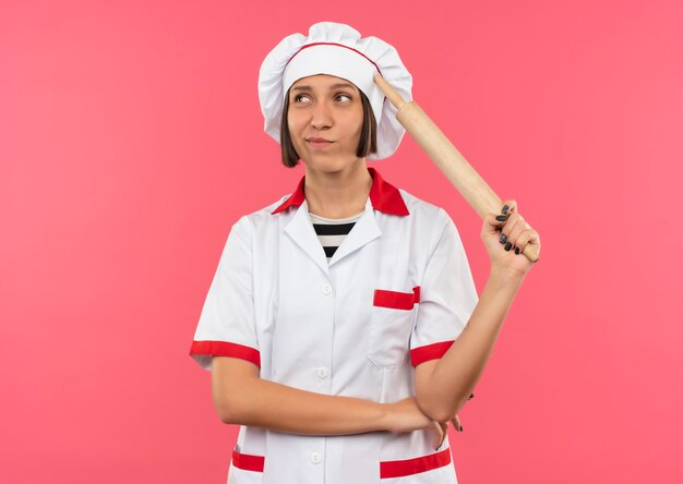 Giovane cuoco femminile premuroso in uniforme del cuoco unico che tiene il mattarello e che mette la mano sotto il gomito che esamina il lato isolato sulla parete rosa