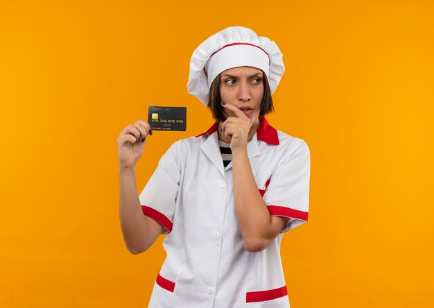 Giovane cuoco femminile premuroso in uniforme del cuoco unico che tiene la carta di credito che esamina il lato e che mette la mano sul mento isolato sulla parete arancione